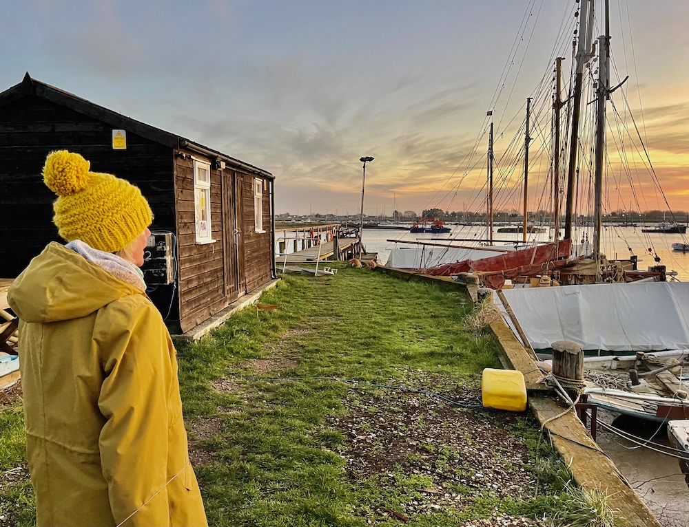 Chrissie Richards in yellow coat and hat standing outside shed in Brightlingsea